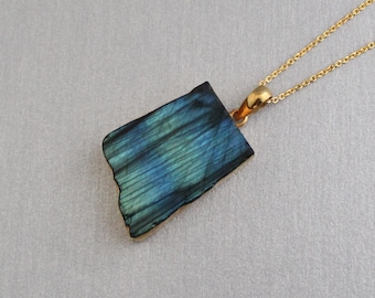 Puede incluir: Un colgante rectangular de labradorita con tonos azules y verdes iridiscentes, suspendido de una fina cadena de metal dorado. La piedra presenta un borde natural ligeramente irregular y una superficie pulida que refleja la luz en patrones lineales.