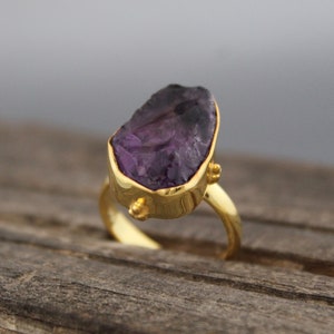 May include: A gold ring featuring a large, irregularly shaped amethyst gemstone. The amethyst is a deep purple colour.