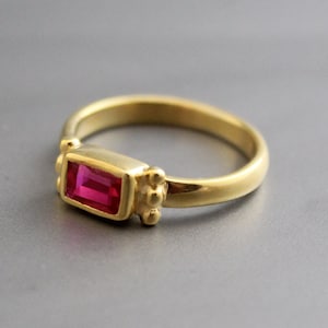 May include: A gold ring with a rectangular red gemstone set in a bezel setting. The ring has a textured band with small beads.