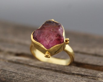 Könnte beinhalten: Ein goldener Ring mit einem großen, facettierten, rosafarbenen Edelstein. Der Ring hat eine einfache Band und der Edelstein ist in einer Zargenfassung gefasst.
