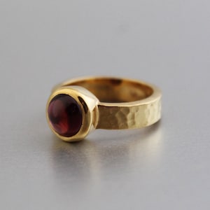 Peut inclure: Une bague en or avec une bande martelée et une pierre précieuse ronde de couleur rouge foncé de forme cabochon.