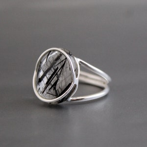 Könnte beinhalten: Ein silberner Ring mit einem schwarzen und klaren Quarzstein. Der Stein ist in einer Zargenfassung gefasst und das Band ist ein einfaches, doppeltes Band.