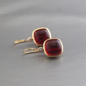 May include: A pair of gold drop earrings featuring square-shaped, deep red cabochon stones. Each earring is accented with a small, clear, round-cut stone set on the gold wire hook, providing a subtle sparkle to the smooth, polished metal design.