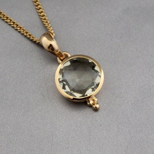 Puede incluir: Un collar de cadena de oro con un colgante que presenta una piedra preciosa redonda de color verde claro engastada en un bisel de oro. El colgante tiene un pequeño detalle decorativo en la parte inferior.