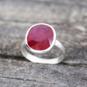 Op de afbeelding: Een zilveren ring met een grote, ovaal geslepen robijn. De robijn heeft een dieprode kleur en is gezet in een eenvoudige, gepolijste zilveren band. De ring wordt gepresenteerd op een getextureerd, grijs oppervlak.