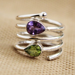 Könnte beinhalten: Ein silberner Ring mit drei übereinanderliegenden Bändern, jedes mit einem birnenförmigen Edelstein besetzt. Der oberste Stein ist Amethyst, der mittlere Stein ist Peridot und der unterste Stein ist ein grüner Edelstein.