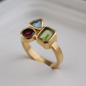 Könnte beinhalten: Ein goldener Ring mit drei Edelsteinen: einem runden roten Stein, einem dreieckigen blauen Stein und einem rechteckigen grünen Stein.