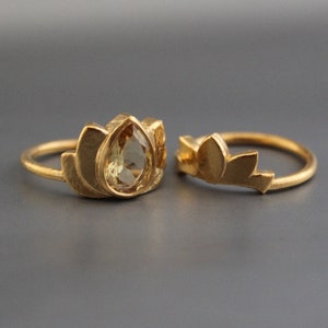 Puede incluir: Dos anillos de oro con un diseño de flor de loto. Un anillo presenta una piedra preciosa amarilla en forma de lágrima.
