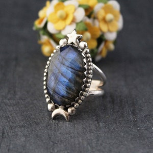 Op de afbeelding: Zilveren ring met een grote, ovale labradoriet edelsteen met blauwe en paarse iriserende kleuren. De ring heeft een decoratieve rand met een ster- en maanontwerp. Geplaatst tegen een donkere achtergrond met gele bloemen.