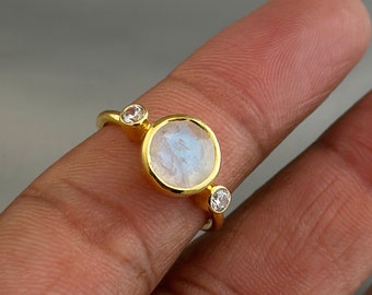 Puede incluir: Un anillo de oro con una gran piedra de luna redonda y dos piedras redondas transparentes más pequeñas. El anillo está en un dedo.