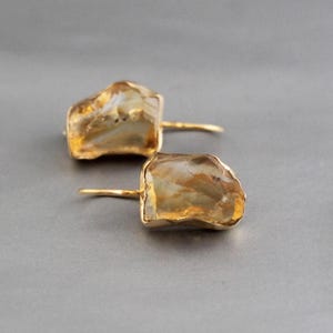Könnte beinhalten: Ein Paar goldfarbene Ohrringe mit unregelmäßigen, hellfarbigen Steineinfassungen. Die Steine haben ein natürliches, organisches Aussehen mit Farb- und Texturvariationen. Die Ohrringe sind an goldenen Stiften befestigt und schaffen ein schlichtes, aber elegantes Design.