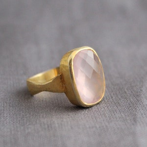 May include: A gold ring with a large, light pink, faceted gemstone. The ring has a simple, hammered band.