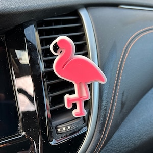 May include: Pink flamingo car air freshener hanging from the air vent of a car. The flamingo is made of plastic and has a white base.