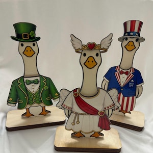 May include: Three wooden goose figurines. One goose wears a green suit and hat with shamrocks, another wears a white dress with a red sash and a heart-shaped crown, and the third wears a blue suit and a top hat with stars.