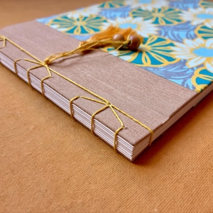 Può includere: Un diario fatto a mano con un motivo floreale blu, giallo e bianco. Il diario ha un dorso in tessuto marrone ed è rilegato con filo dorato. Un piccolo nappa con perline di legno adorna la parte superiore.