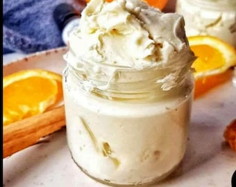 May include: A clear glass jar filled with a thick, off-white whipped cream or body butter. The product has a textured, swirled surface. It is surrounded by fresh orange slices and brown cinnamon sticks, with a whole orange visible in the back.