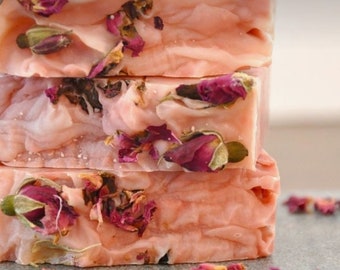 May include: A stack of three rectangular bars of light pink soap. Each bar features a textured surface embedded with dried rosebuds and scattered dark pink rose petals. The soap has a smooth, creamy appearance with visible folds and layers.