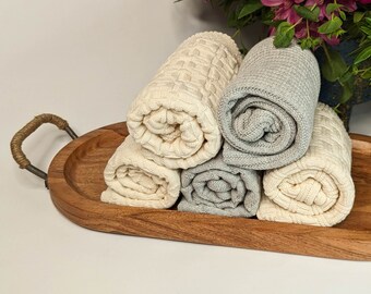 May include: Five rolled up knitted towels in various shades of cream and gray are displayed on a wooden tray with a metal handle.