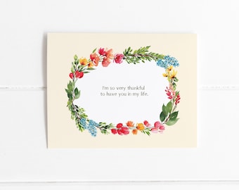 May include: A cream-colored greeting card with a floral wreath border. The wreath features a variety of colorful flowers and green leaves. The text inside the wreath reads, "I'm so very thankful to have you in my life."