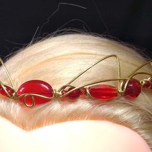 Peut inclure: Une couronne en fil d'or avec des perles de verre rouges. La couronne est conçue avec un style simple et élégant.