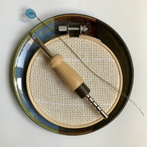 Pode incluir: Um close-up de um aro de bordado de madeira com um tecido branco esticado sobre ele. Uma ferramenta de agulha de metal com uma alça de madeira está sobre o tecido. Uma conta azul está presa a um fio fino que também está sobre o tecido.