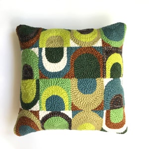 Puede incluir: Una almohada cuadrada con un patrón geométrico de círculos y arcos verdes, marrones y azules. La almohada está hecha de una tela texturizada y tiene un fondo blanco.
