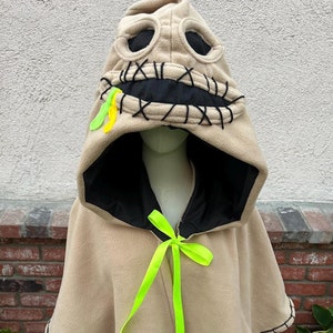 May include: A tan fleece hooded cape with a stitched-on face, black eyes, and a stitched-on mouth. The hood is lined with black fleece. The cape has a green ribbon tied at the neck.
