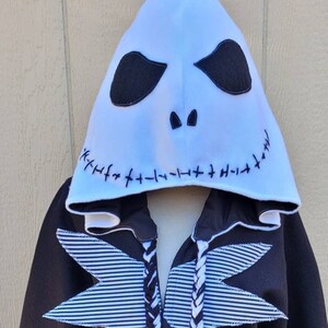 May include: Black hooded sweatshirt with a white Jack Skellington face on the hood. The sweatshirt has white and black striped fabric on the front and black and white braided drawstrings.