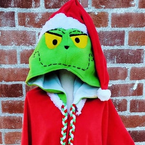 May include: Red hooded sweatshirt with a green Grinch face on the hood. The hood has a white trim and a red Santa hat on top. The sweatshirt has white fur trim on the sleeves and a green and white braided cord.