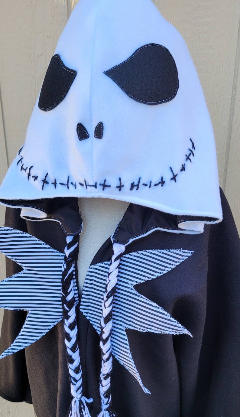 May include: Black and white striped fabric wings with a white hooded sweatshirt featuring a black stitched Jack Skellington face. The hood has a white fleece lining.