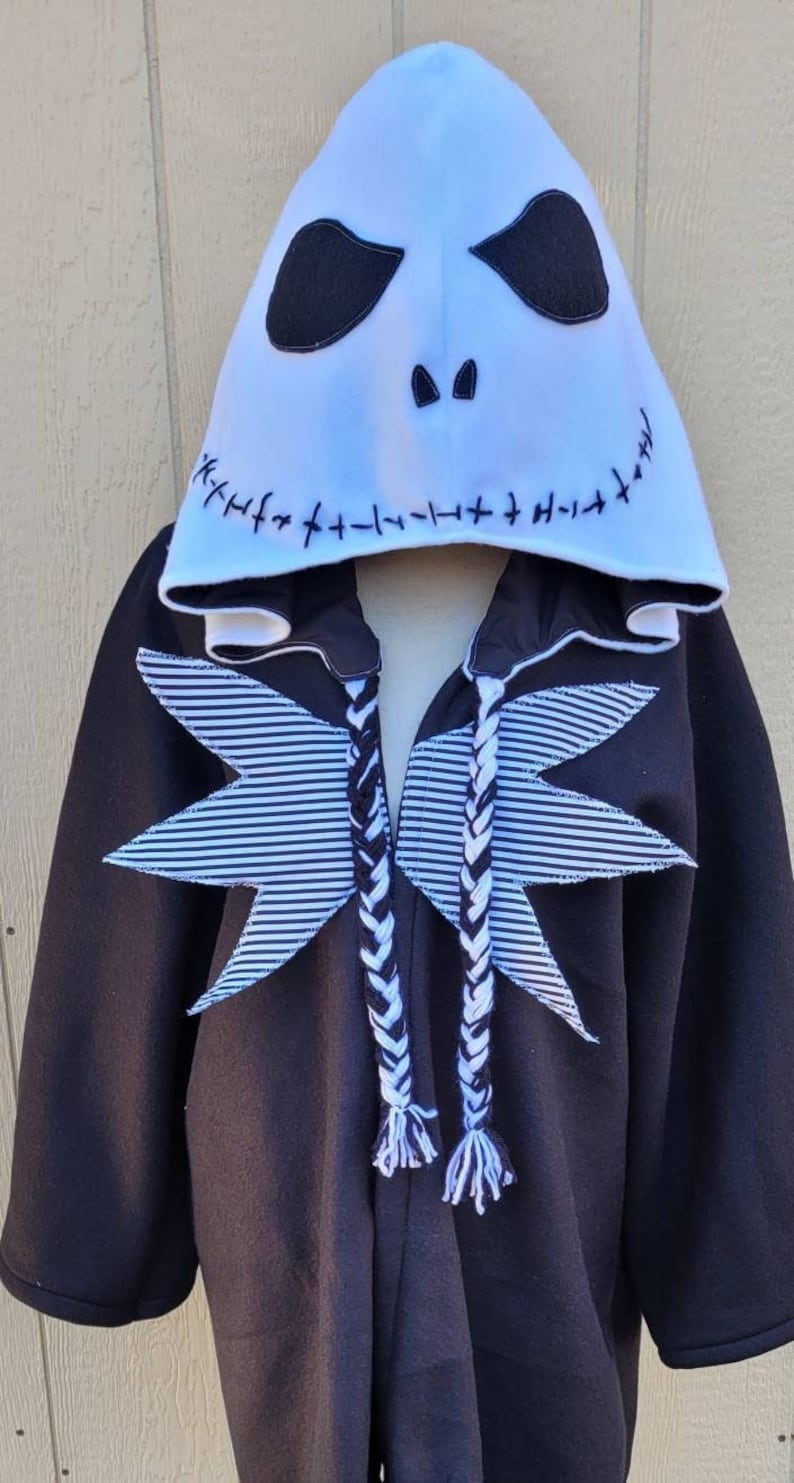 May include: Black hooded sweatshirt with a white Jack Skellington face on the hood. The sweatshirt has white and black striped fabric on the front and black and white braided drawstrings.
