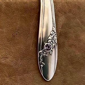 May include: A silver-toned metal bookmark shaped like a curved spoon handle, featuring an embossed floral pattern along one side and a single, round, purple faceted stone set into the center of the decorative design.