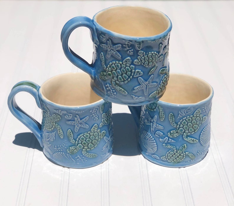 Puede incluir: Tres tazas de cer&aacute;mica azul con un dise&ntilde;o de tortuga marina y estrella de mar. Las tazas est&aacute;n apiladas una encima de la otra.