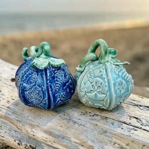 May include: Two ceramic pumpkin-shaped ornaments featuring embossed seashell, starfish, and sand dollar patterns. One pumpkin is a deep royal blue, while the other is a lighter, pale turquoise blue. Both have sculpted stems and leaves on top.