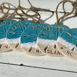 May include: A collection of ceramic sand dollar ornaments with a coastal theme. Each ornament is a different shade of blue and white, with text indicating coastal locations. They are strung with natural twine.