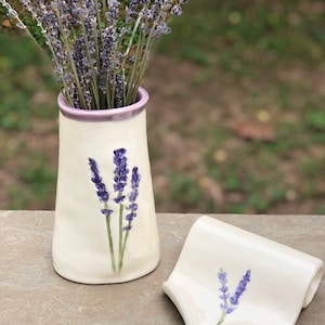 May include: Two white ceramic vases with lavender flower designs. The taller vase has a purple rim and is filled with dried lavender. The smaller vase is shaped like a rolled-up piece of paper.