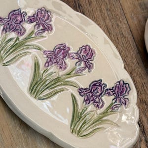 May include: Cream-colored ceramic serving platter with a scalloped edge. The platter is decorated with raised purple iris flowers and green leaves. The flowers are arranged in a naturalistic pattern across the surface.