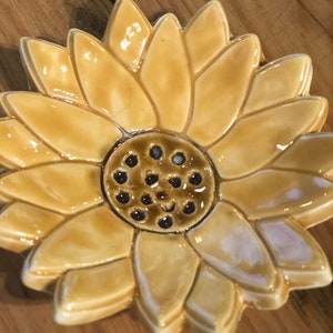 May include: A ceramic sunflower dish with a yellow glaze and a brown center. The center has a small smiley face.