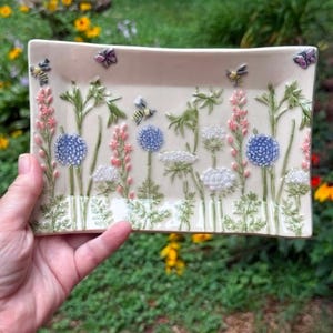 May include: A rectangular ceramic tray with a cream-colored base featuring a raised, three-dimensional garden scene. The design includes green stems, pink and blue floral clusters, white Queen Anne's lace, small bees, and purple butterflies.