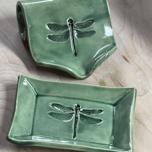 May include: Two green ceramic soap dishes. One is rectangular with a raised edge, and the other has a curved handle. Both feature a dragonfly design in the center. The dishes are on a light wood surface.