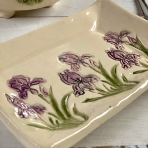 May include: A white ceramic dish with a raised design of purple iris flowers and green leaves. The dish has a slightly rounded rectangular shape.