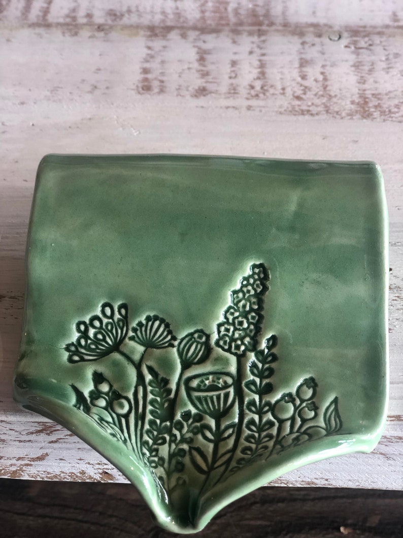May include: A green ceramic dish with a raised design of stylized flowers and leaves. The dish has a wavy edge and a slightly curved shape.