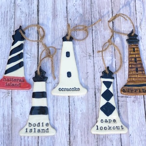 May include: Five ceramic lighthouse ornaments with different designs and names. The ornaments are white with black and red stripes, black and white stripes, black and white diamonds, white with black dots, and brown with black stripes. The names of the lighthouses are Hatteras Island, Bodie Island, Cape Lookout, Ocracoke, and Currituck.
