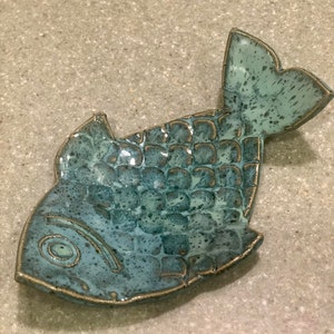 May include: A teal blue ceramic fish dish with a textured surface. The dish is shaped like a fish with a tail fin and a raised eye.