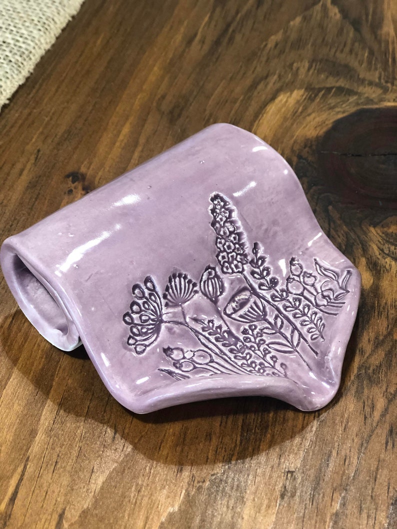 May include: A purple ceramic dish with a floral design. The dish is shaped like a rolled-up piece of fabric.