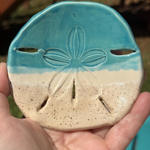 May include: A ceramic sand dollar-shaped dish with a two-tone design. The top half is a glossy turquoise, transitioning to a sandy beige at the bottom. The dish features a detailed, embossed floral pattern and several small holes.