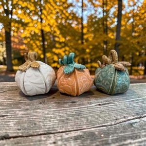 May include: Three ceramic pumpkin figurines featuring embossed leaf patterns. The left pumpkin is white with a tan stem, the center is orange with a green stem, and the right is teal with a tan stem. Each piece has a glossy finish and curved stems.