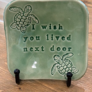 May include: A square, seafoam green ceramic dish with the words "I wish you lived next door" embossed on the surface. Two turtle designs are also embossed on the dish. The dish is displayed on a wooden surface.