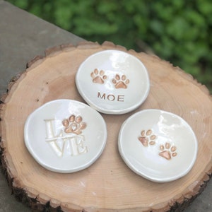May include: Three white ceramic ring dishes with gold paw prints and personalized text. The dishes are on a wooden surface. The text on the dishes reads "LOVE", "Moe", and two paw prints.