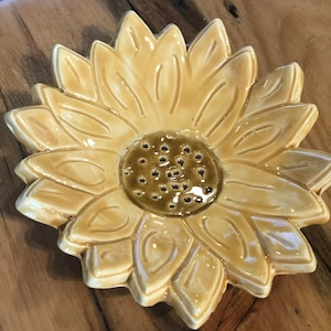May include: A ceramic sunflower dish with a yellow glaze and a brown center. The center has small holes for drainage.